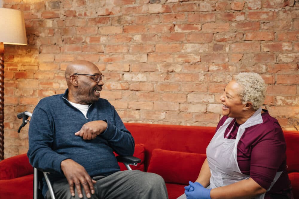 A smiling healthcare worker talks with an older man in a wheelchair, both sitting in front of a brick wall background. - Home Instead