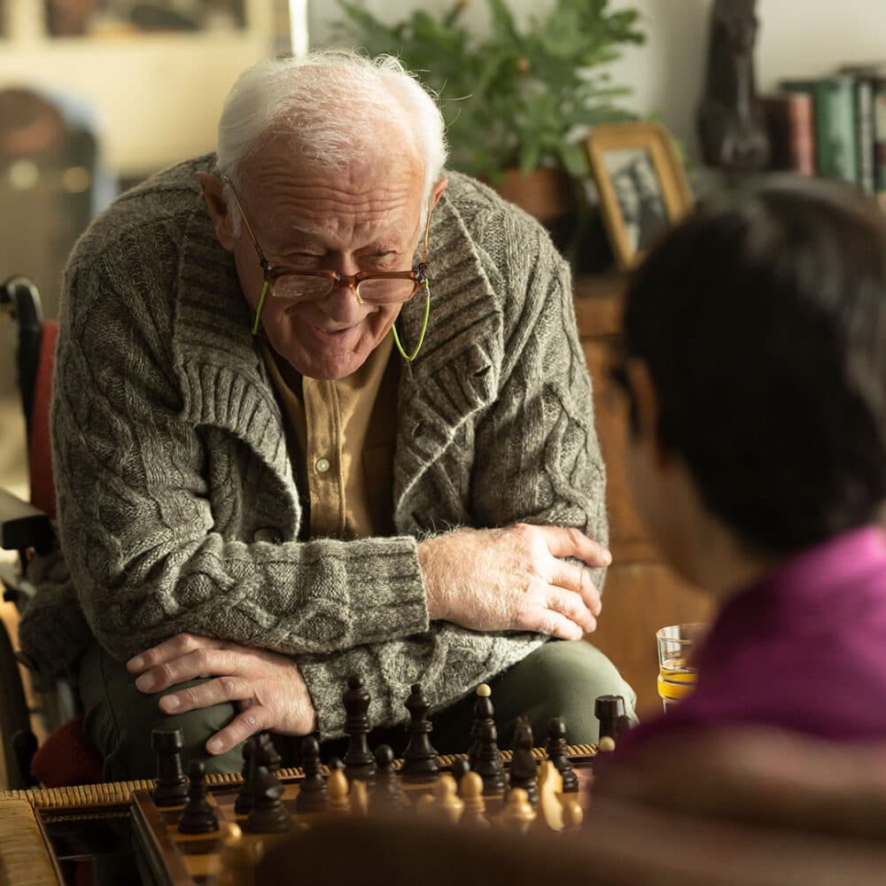 An elderly man in a gray sweater plays chess with a younger person, smiling and leaning forward over the chessboard. - Home Instead