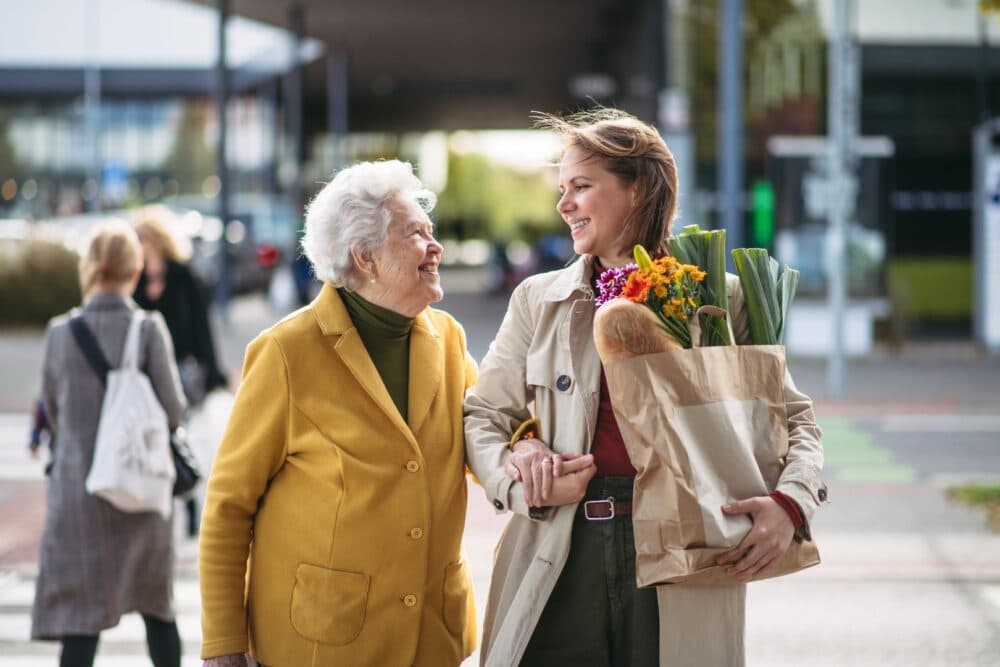 An elderly woman and a young woman smiling and walking together, with the young woman holding a paper bag of groceries. - Home Instead