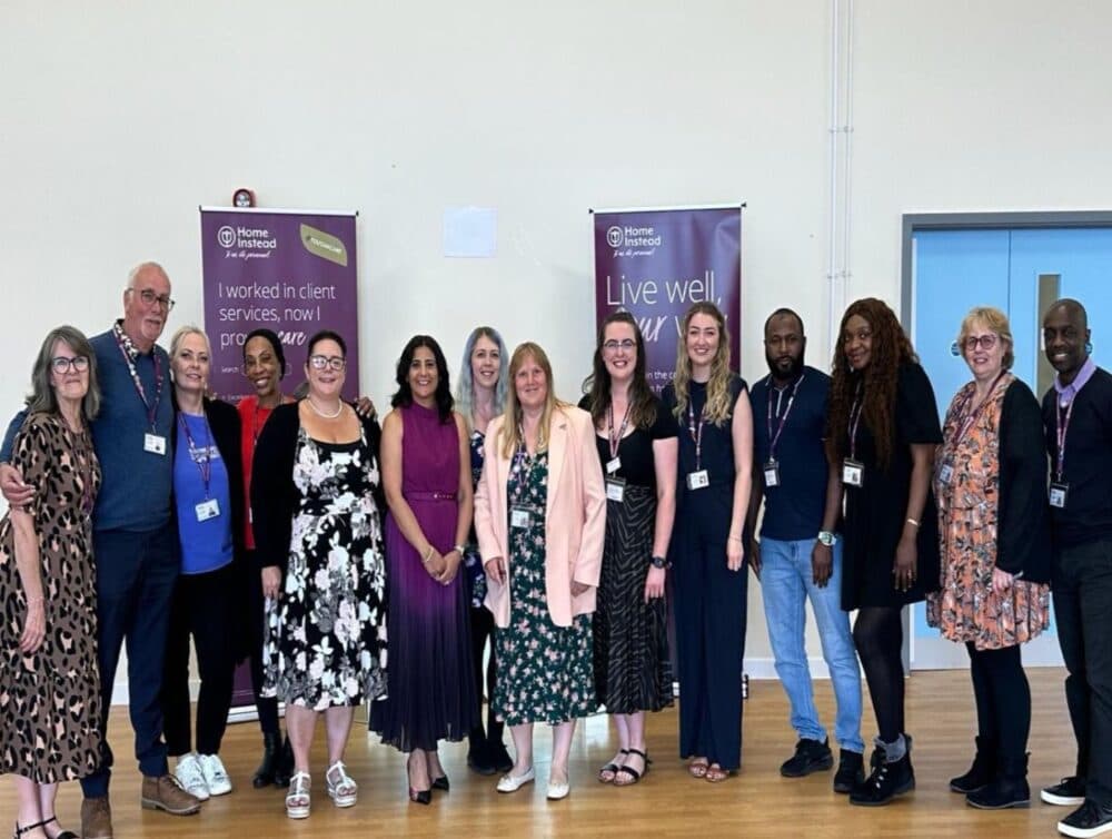 A group of people standing in a row indoors, with banners about client services and living well in the background. - Home Instead