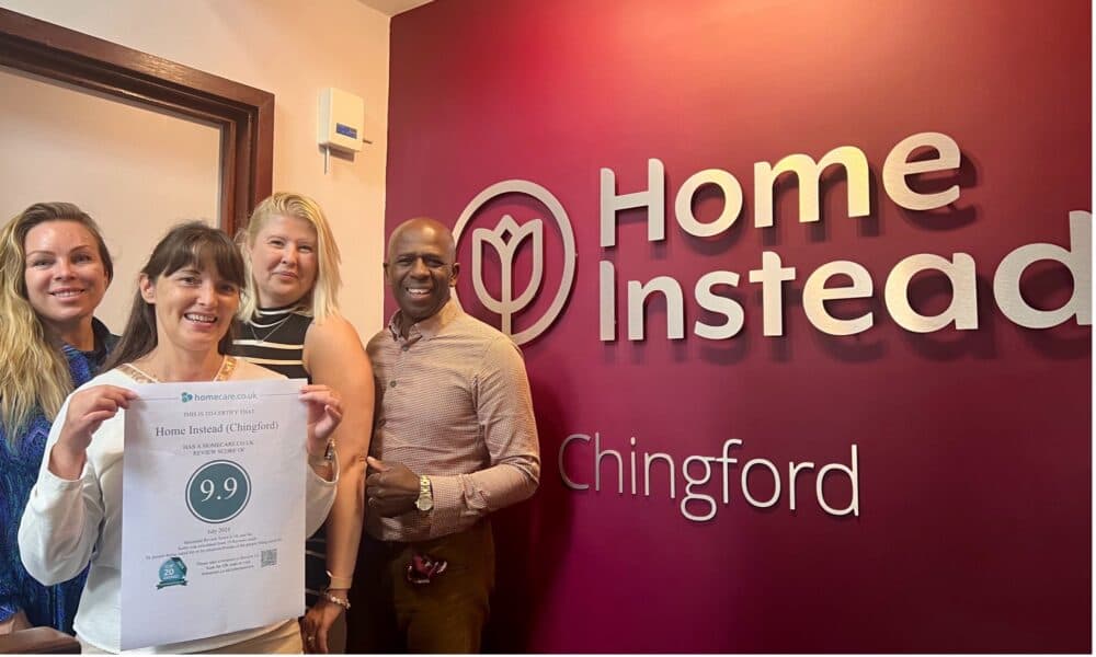 Four people smiling, with one holding a 9.9 rating sign in front of a wall displaying the Home Instead Chingford logo. - Home Instead