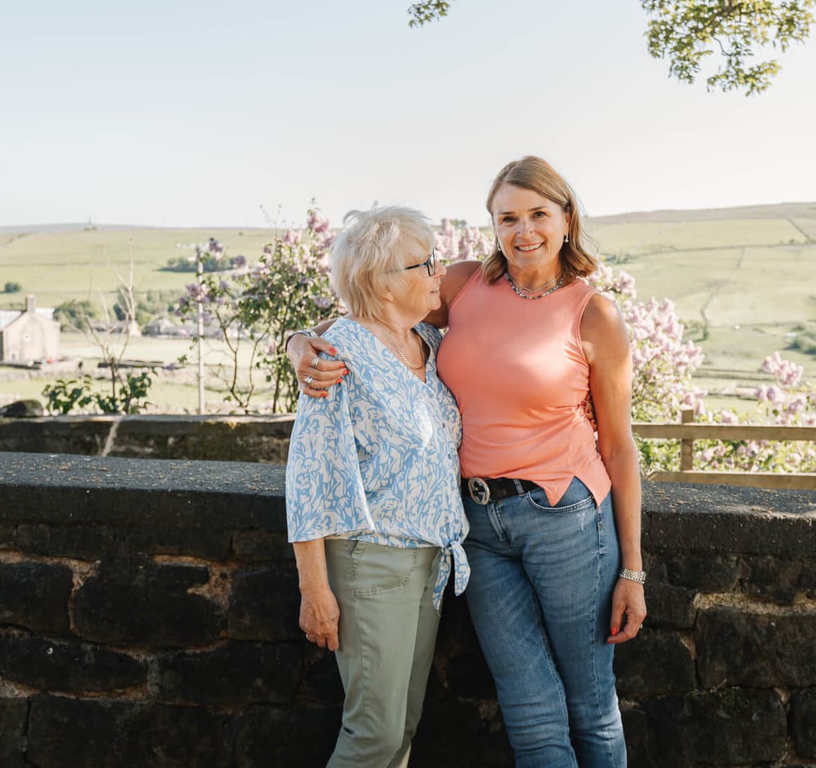 Two women standing outdoors, one in a blue blouse, and the other in a pink top, smiling and posing with countryside behind. - Home Instead