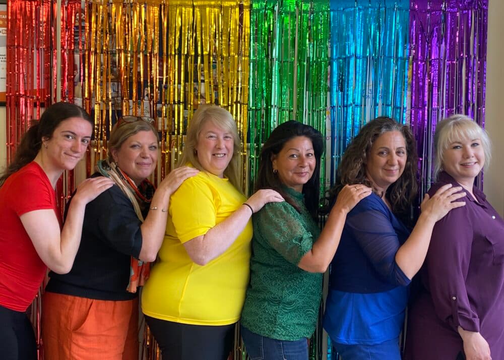 A group of six women stands in a line with hands on each other's shoulders against a rainbow fringe background. - Home Instead