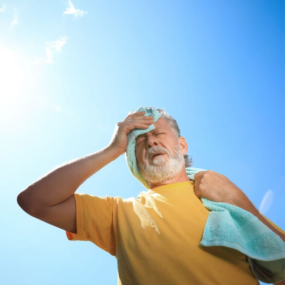 Elderly man in a yellow shirt wiping sweat from his forehead with a towel on a sunny day. - Home Instead