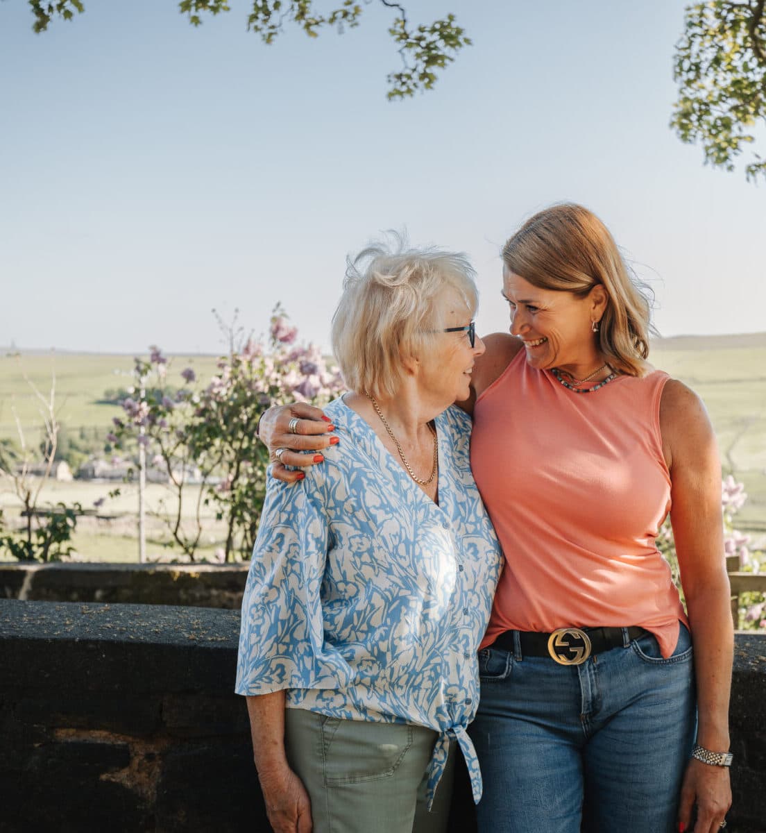 Two women, one elderly in a floral shirt and one middle-aged in a coral tank top, embrace outdoors on a sunny day. - Home Instead