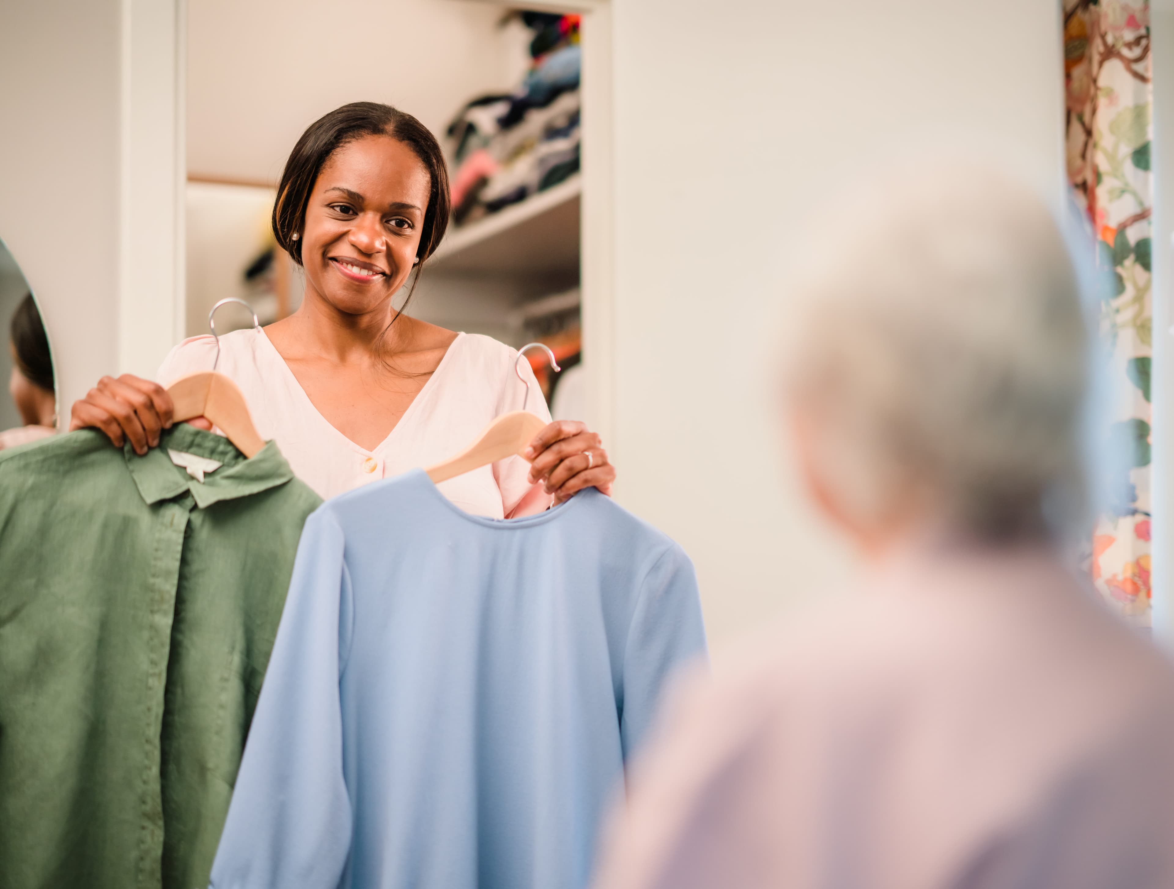 Woman holding a green shirt and a blue shirt on hangers, smiling, while an older person looks on in a bedroom. - Home Instead
