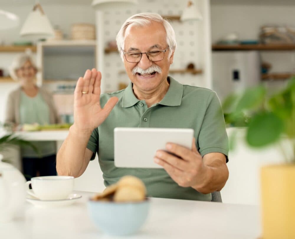 Elderly man smiling and waving at tablet during video call, sitting at table with coffee and snacks. - Home Instead
