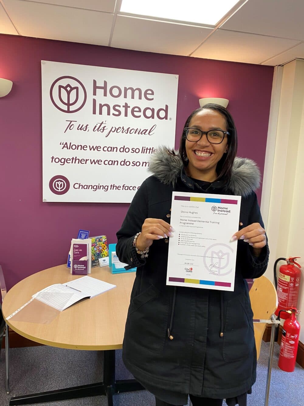 A person smiling and holding a certificate in an office with a Home Instead sign on the wall behind them. - Home Instead