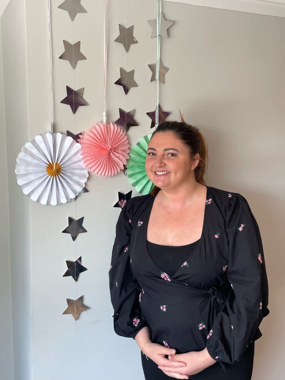 A smiling woman stands in front of a wall with hanging star decorations and colorful paper rosettes. - Home Instead