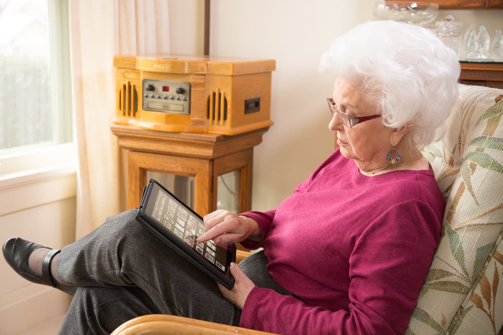 Elderly woman with white hair and glasses, seated in a chair, uses a tablet. Vintage radio and window in background. - Home Instead