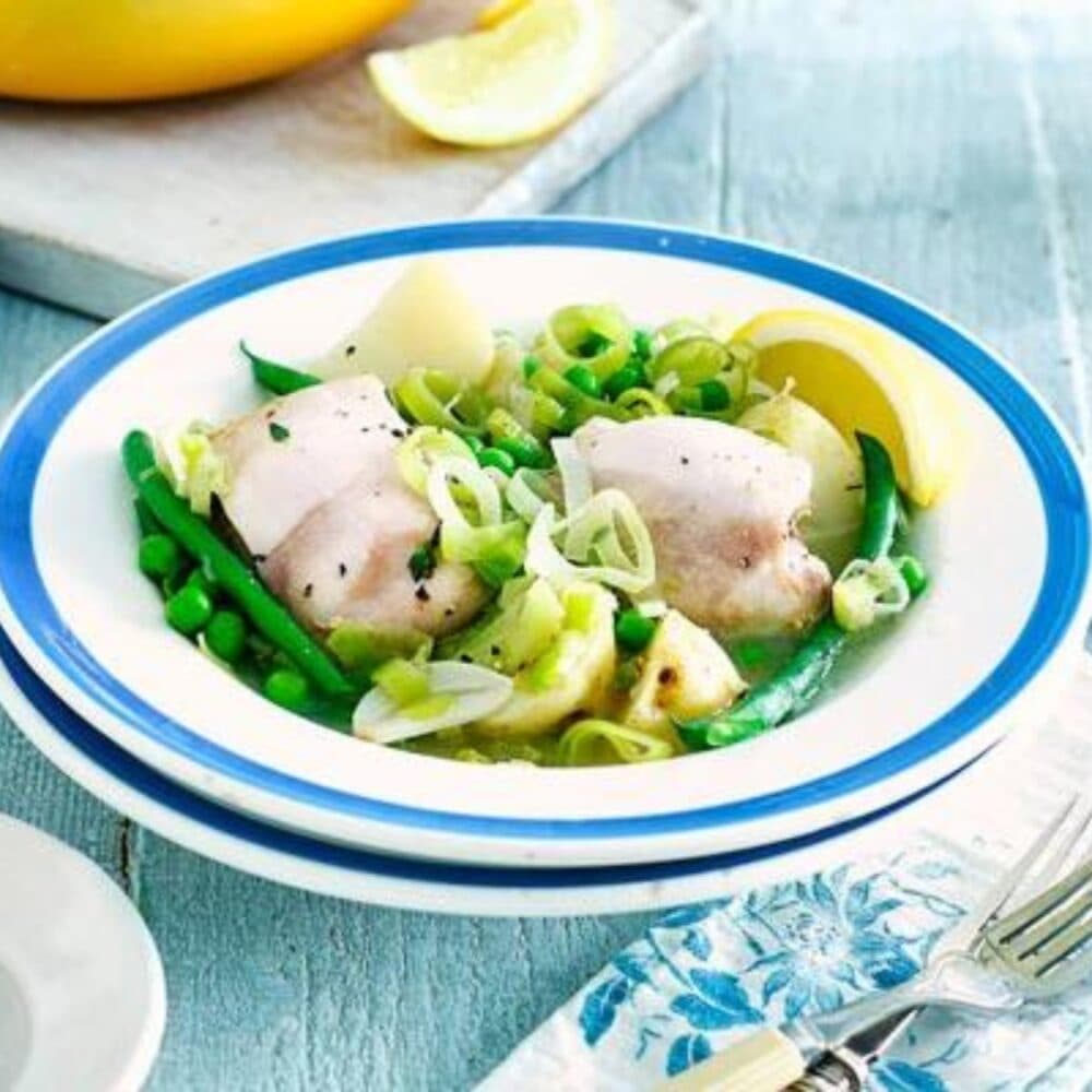 A bowl of cooked chicken pieces with green beans, peas, and leek, garnished with lemon slices on a blue table. - Home Instead Renfrewshire and Barrhead home care services - meal preparation.