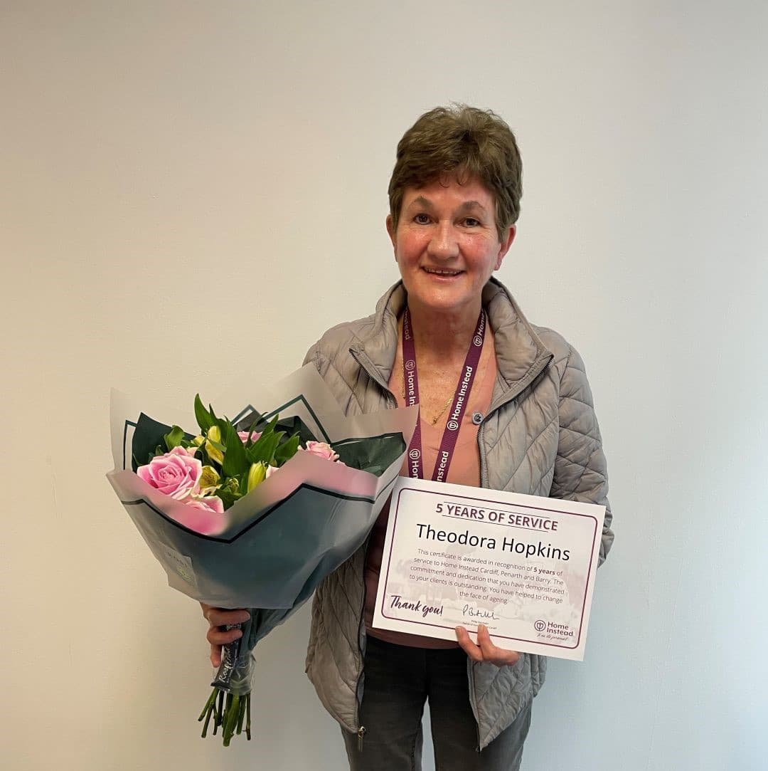 Person holding a certificate and a bouquet of flowers, smiling against a plain background. - Home Instead