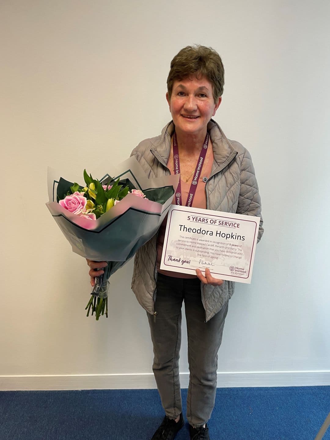 A person holds a bouquet of flowers and a "5 Years of Service" certificate while smiling in front of a plain background. - Home Instead
