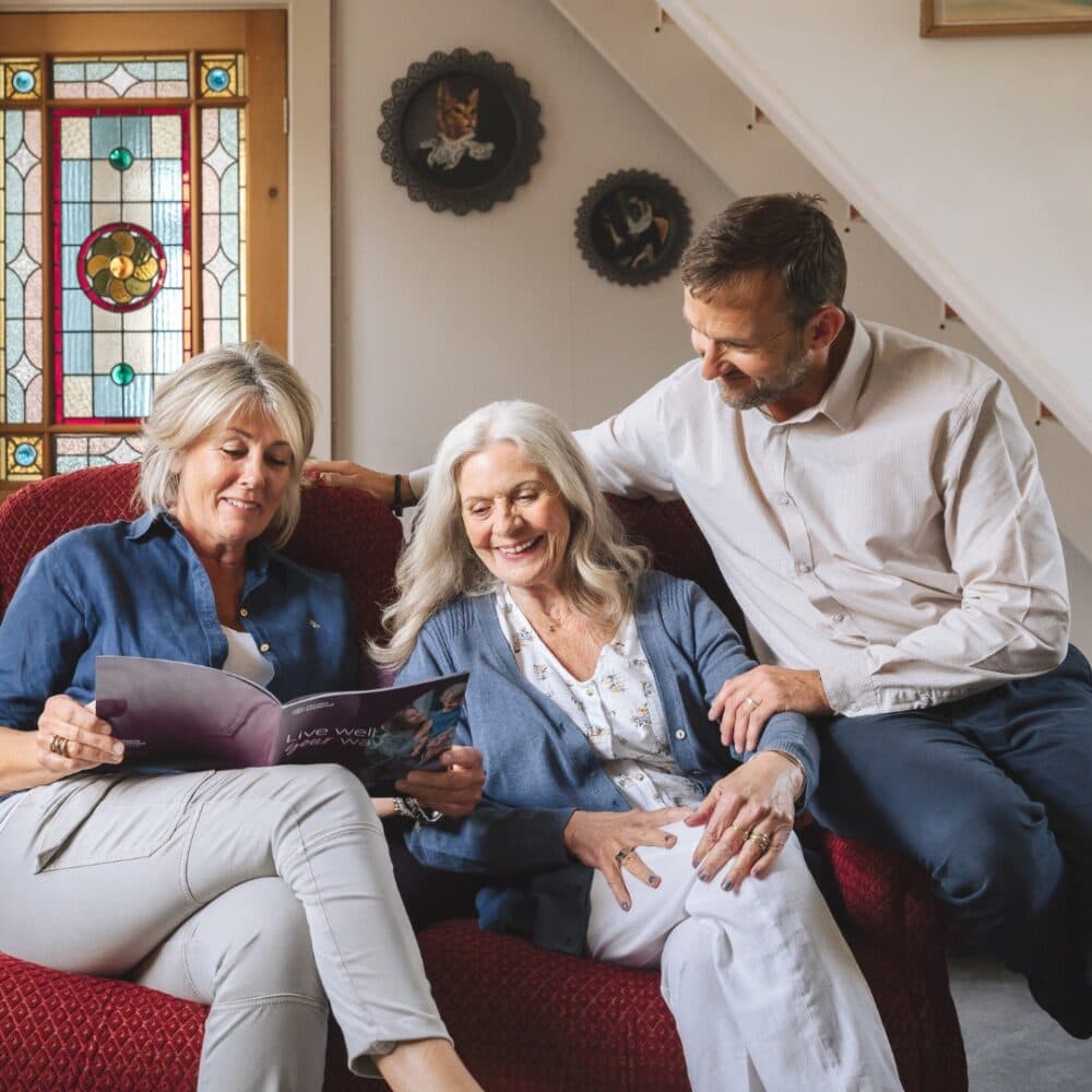 Three people sitting on a red couch, smiling and looking at a magazine in a cozy room with stained glass windows. - Home Instead