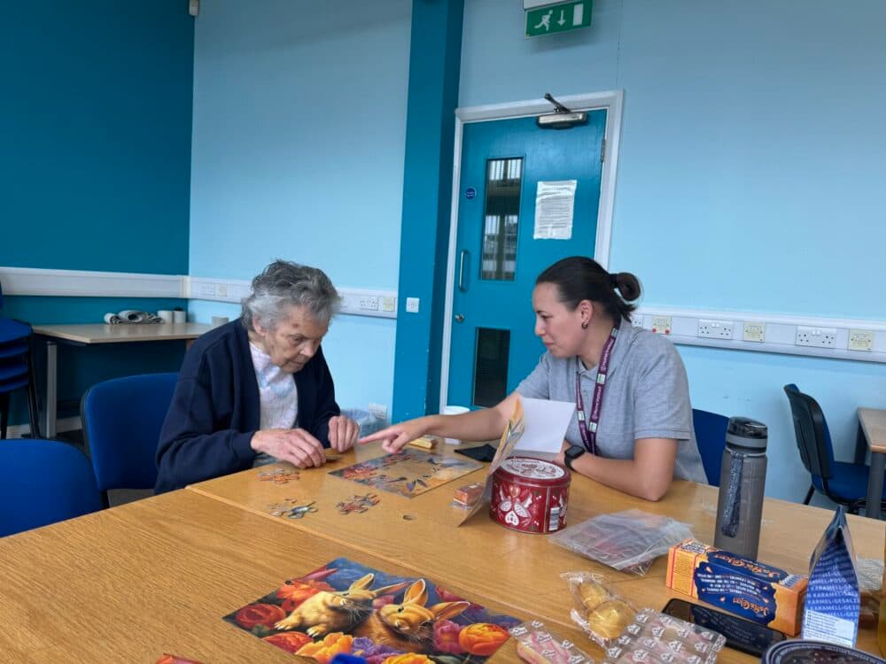 An elderly woman and a younger woman assemble a puzzle together at a table in a brightly lit room. - Home Instead