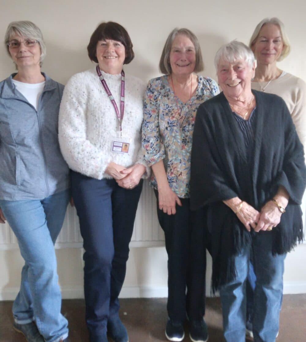 Five older women stand together indoors, smiling at the camera in casual clothing. - Home Instead