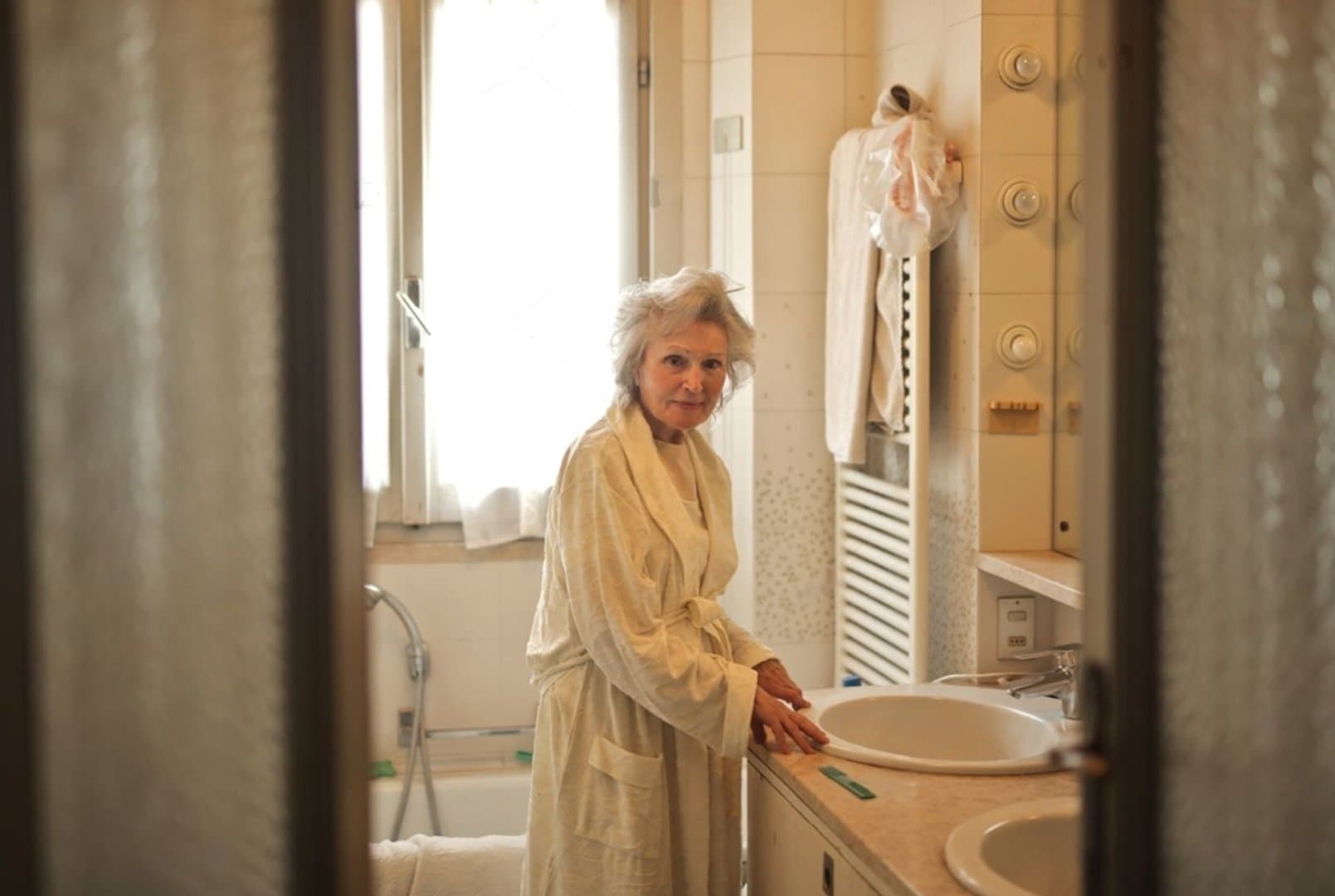 An elderly woman wearing a robe stands in a bathroom, leaning on the sink and looking toward the camera. - Home Instead