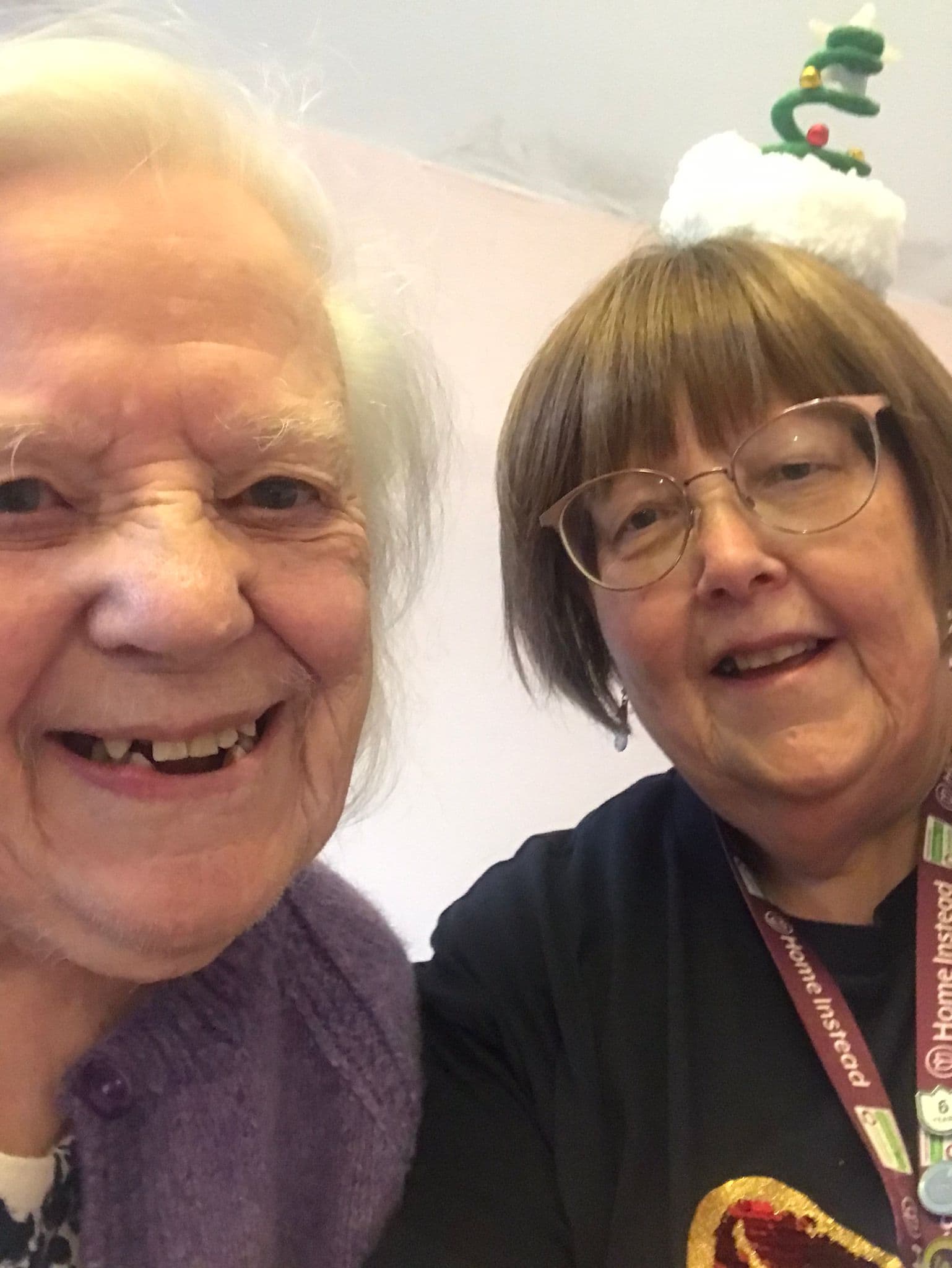 Two elderly smiling women; one wears a Christmas-themed headband and glasses, the other has white hair and wears a purple sweater. - Home Instead