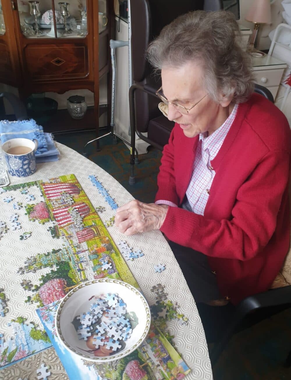 An elderly woman with glasses and a red cardigan works on a colorful jigsaw puzzle at a table, smiling. - Home Instead