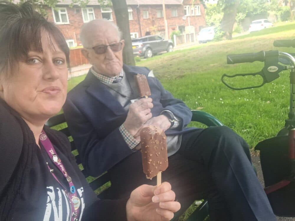 Two people sitting on a bench eating ice cream bars, one holding a walking aid. Green grassy area and houses in the background. - Home Instead