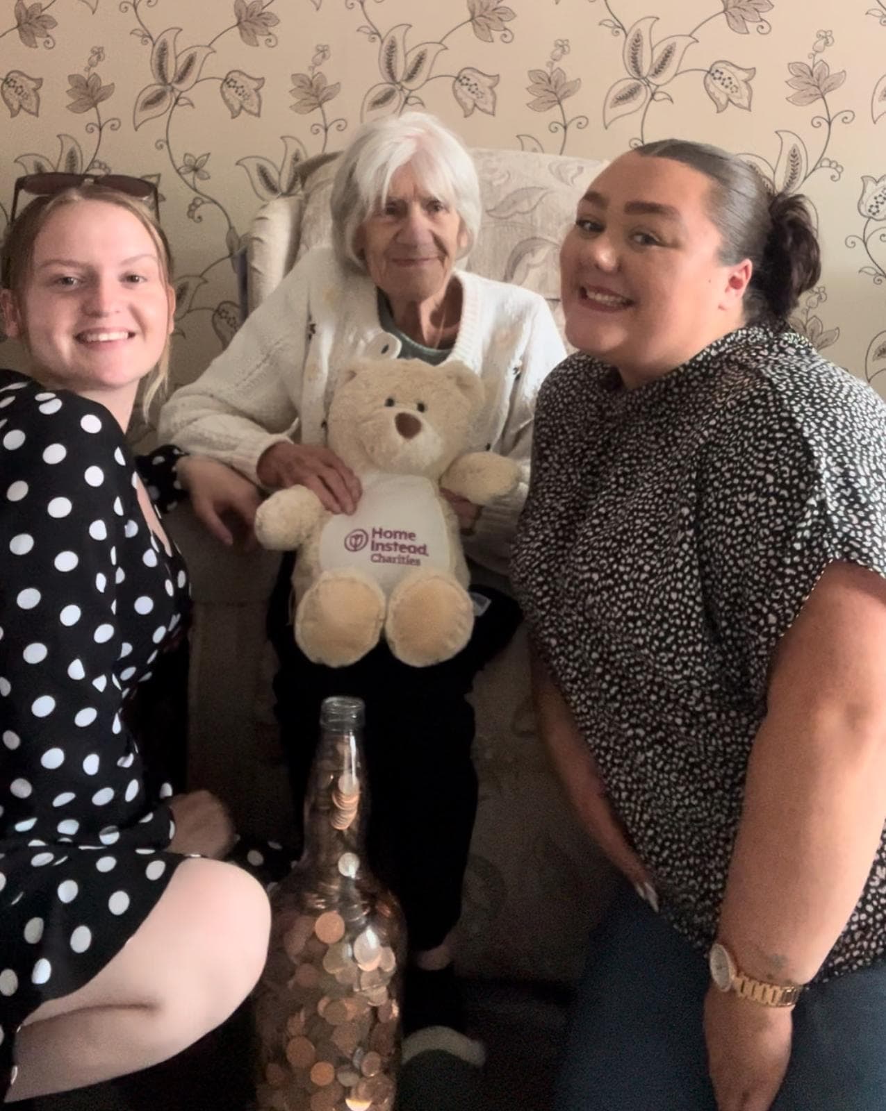 Three women pose with a teddy bear and a large jar filled with coins. The background has floral wallpaper. - Home Instead