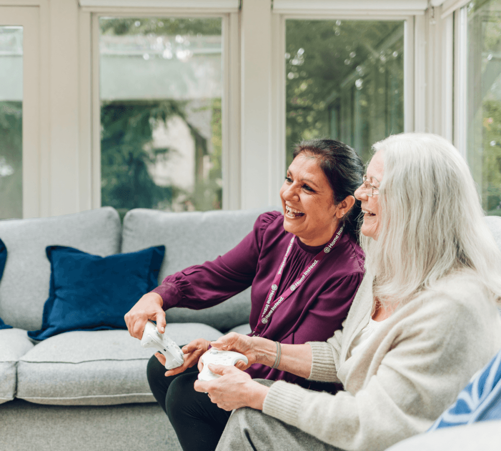 Two women smiling and playing video games on a couch in a bright, sunlit room with blue and gray decor. - Home Instead