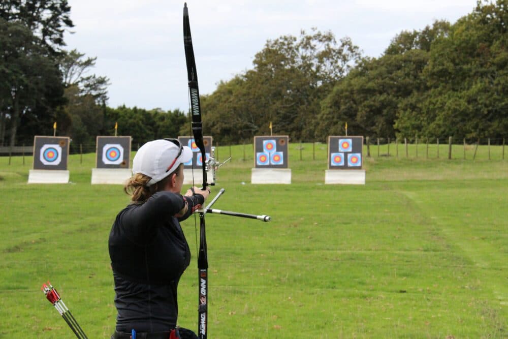 Person aiming a bow and arrow at multiple targets set up in an open grassy field with trees in the background. - Home Instead