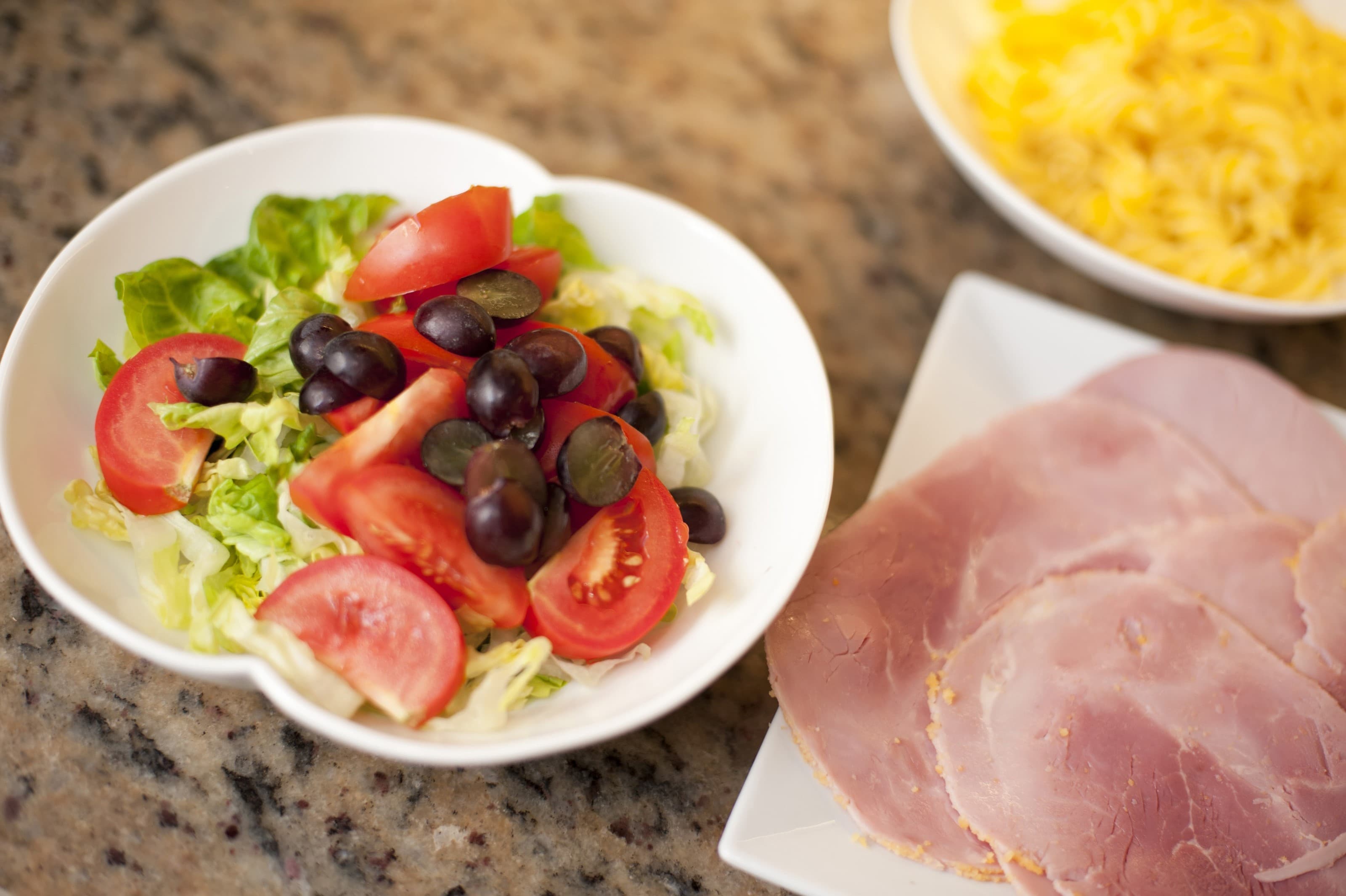 A bowl of salad with lettuce, tomatoes, and grapes next to a plate of sliced ham on a granite countertop. - Home Instead Renfrewshire and Barrhead Care at Home services