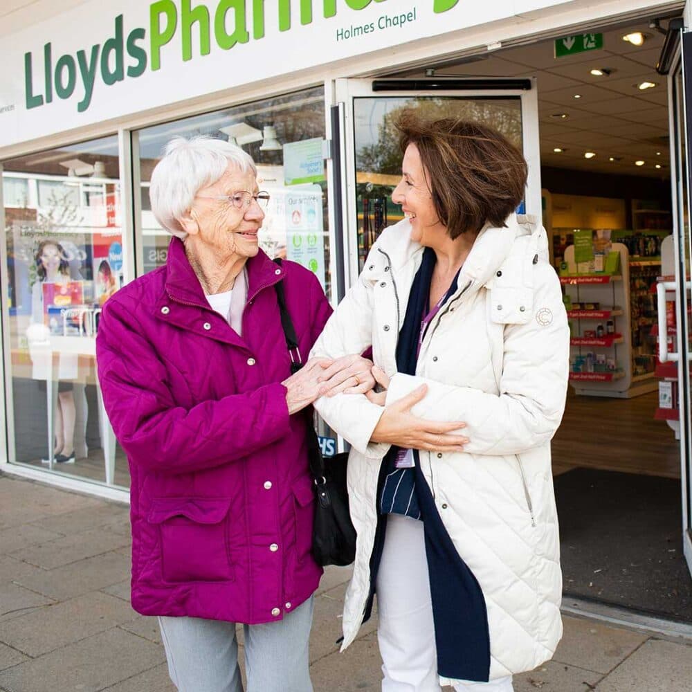 Two women smiling and chatting outside a LloydsPharmacy store, one in a purple coat and the other in a white coat. - Home Instead