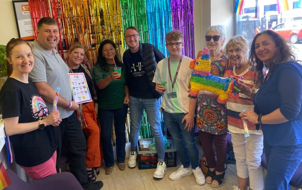 A group of people smiling and posing with a rainbow piñata and colorful drinks in front of a rainbow backdrop. - Home Instead