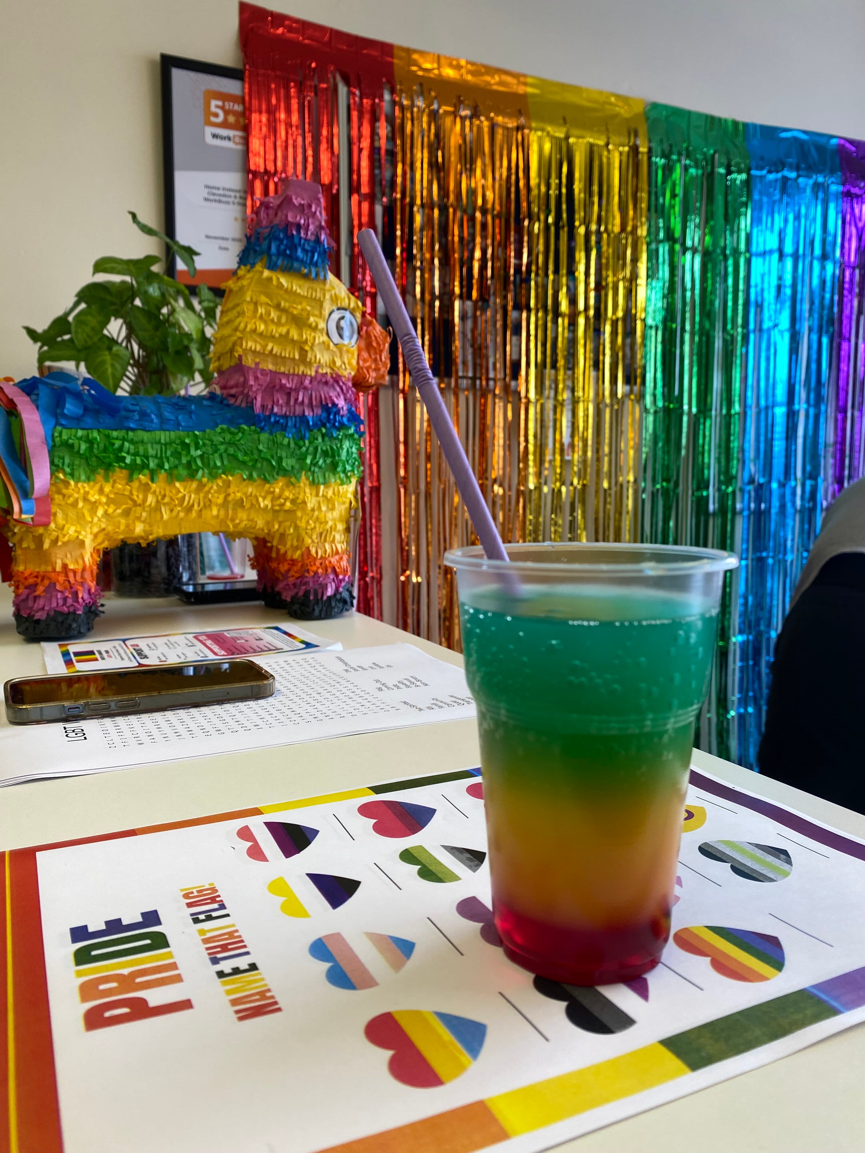 A colorful layered drink in a plastic cup with a straw sits on a table. Pride-themed decorations are in the background. - Home Instead