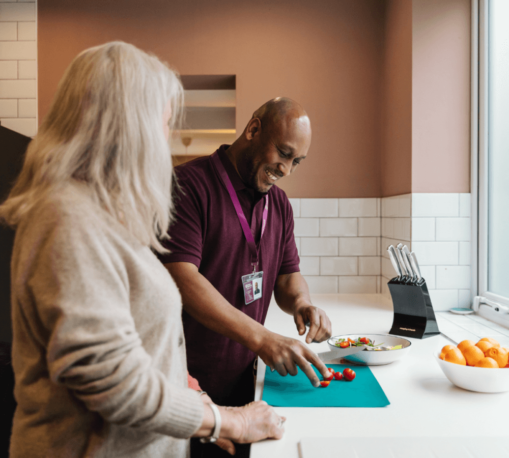 A man chops vegetables on a cutting board while a woman watches in a bright kitchen. - Home Instead