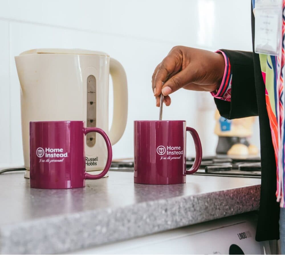 A person stirs a drink in a maroon "Home Instead" mug next to another identical mug on a kitchen counter with a kettle. - Home Instead Southampton