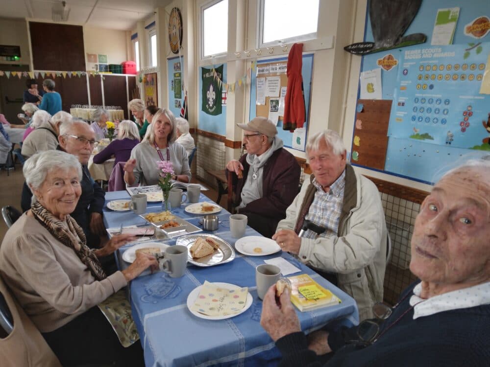 Elderly people sit around a table, enjoying tea and snacks in a bright, decorated community room. - Home Instead