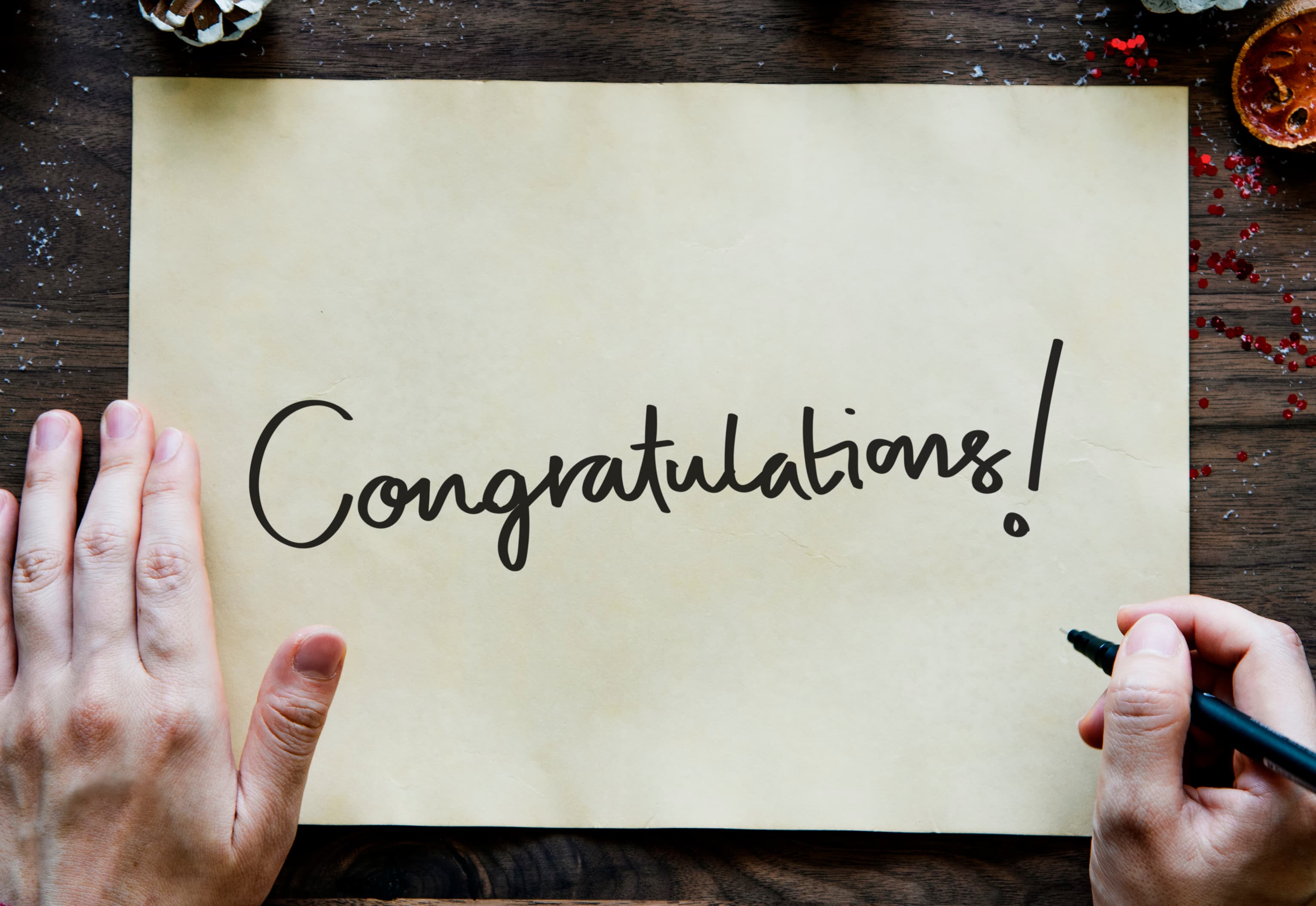 Hands holding paper with "Congratulations!" written on it in cursive, with pen and festive items around. - Home Instead
