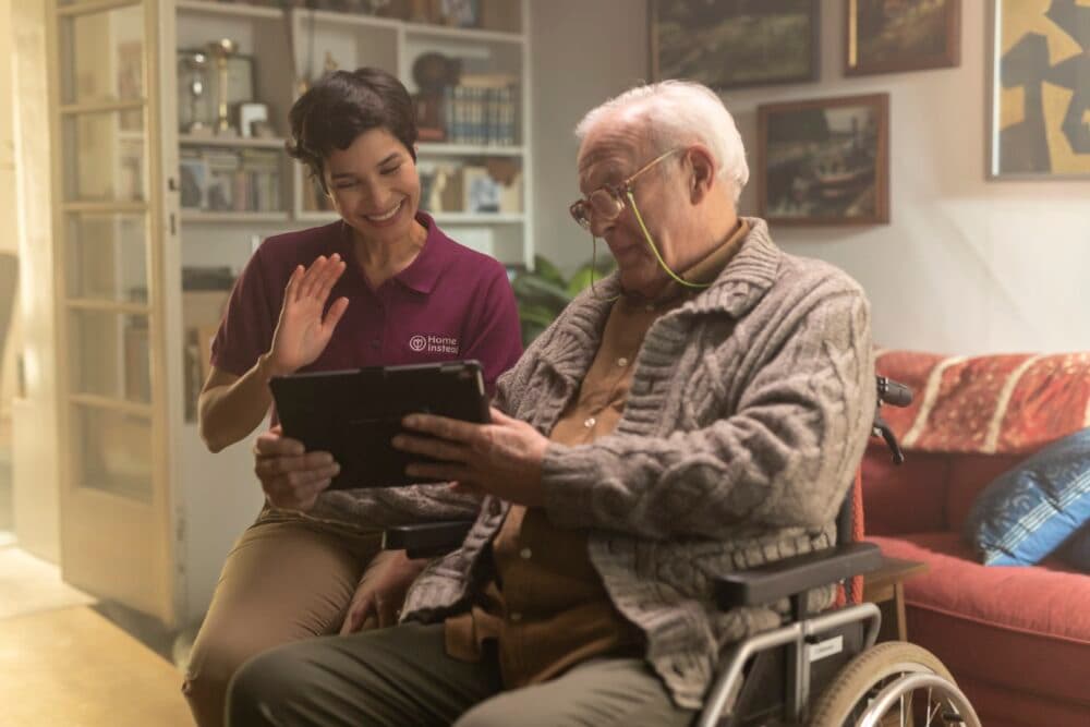 A caregiver smiles and waves, sitting beside an elderly man in a wheelchair who is holding a tablet. - Home Instead