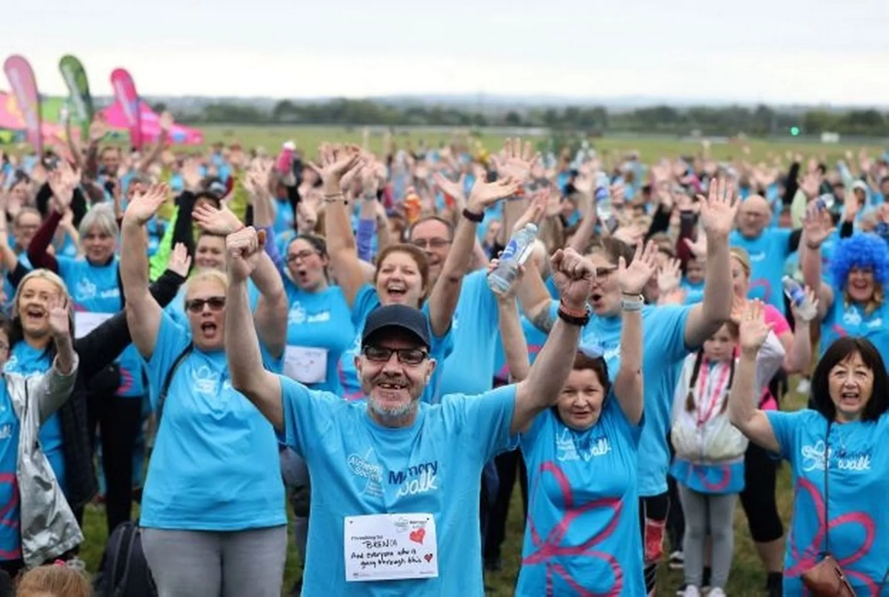 A large group of people wearing blue shirts raise their hands and smile outdoors at a fun event. - Home Instead