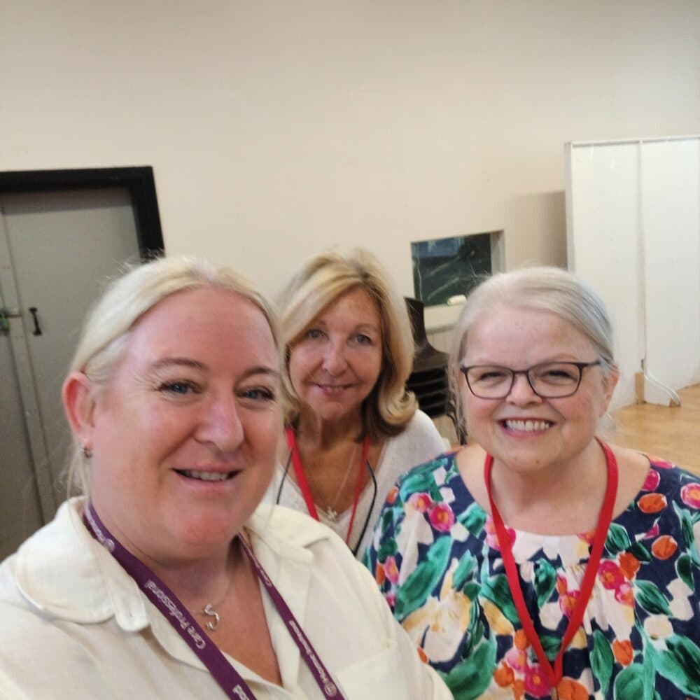 Three women smiling, standing together, one wearing glasses, all with light-colored hair, in a room with a neutral background. - Home Instead