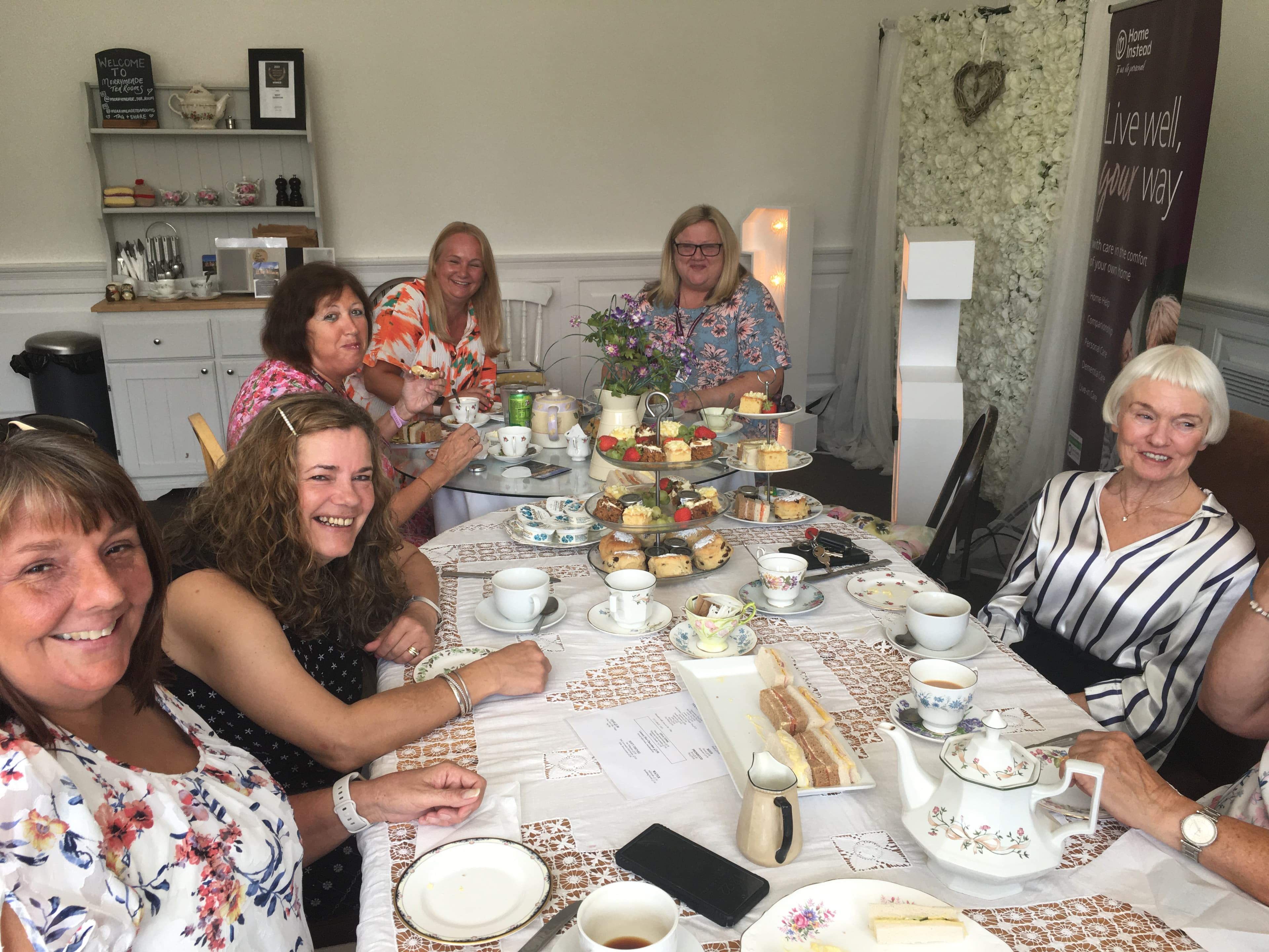 A group of women smiling and enjoying afternoon tea together at a table adorned with various tea sets and treats. - Home Instead