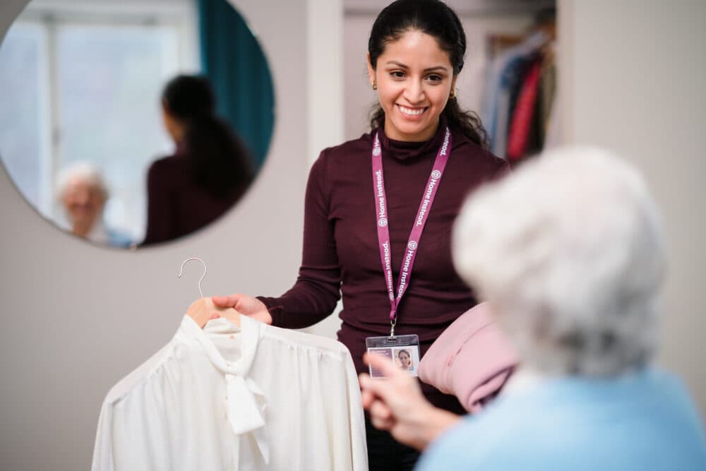 A caregiver smiling, holding a white blouse, while assisting an elderly person in a room with a round mirror. - Home Instead