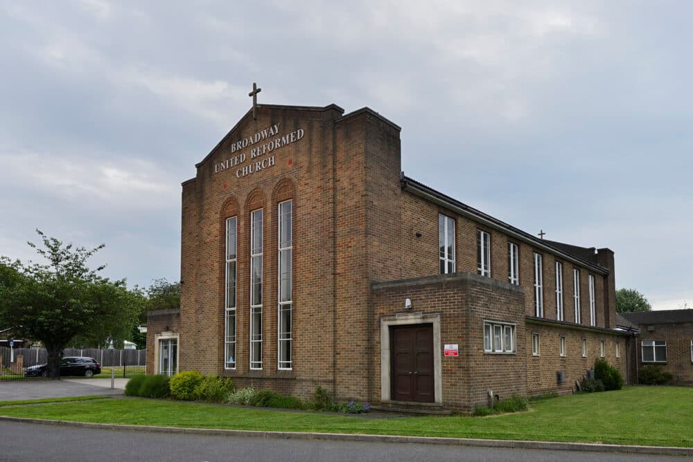Brick church building with tall windows, arched entrance, and a cross on top. The sign reads "Broadway United Reformed Church. - Home Instead