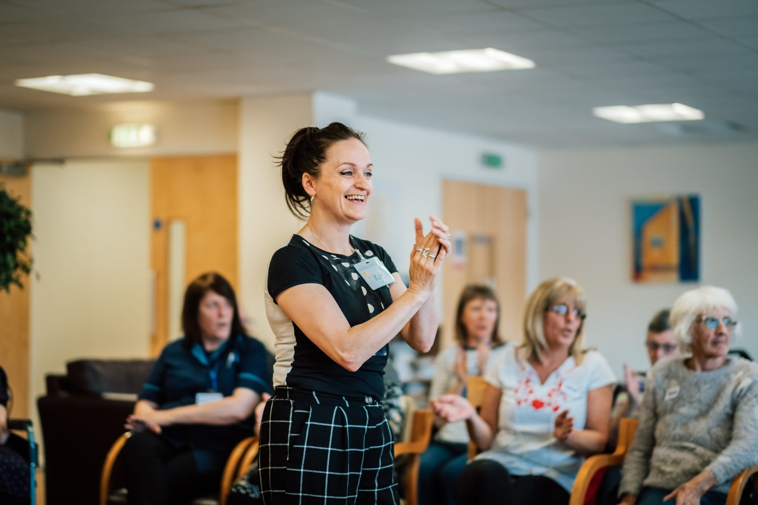 A woman stands clapping in a room with seated people who are watching her attentively. - Home Instead