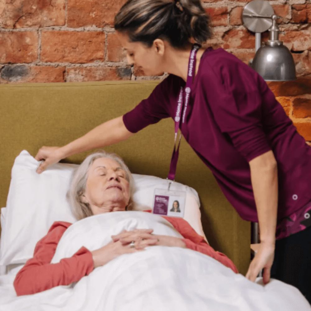 A caregiver in a maroon top assists an elderly woman lying in bed, adjusting her pillow in a cozy room with brick walls. - Home Instead