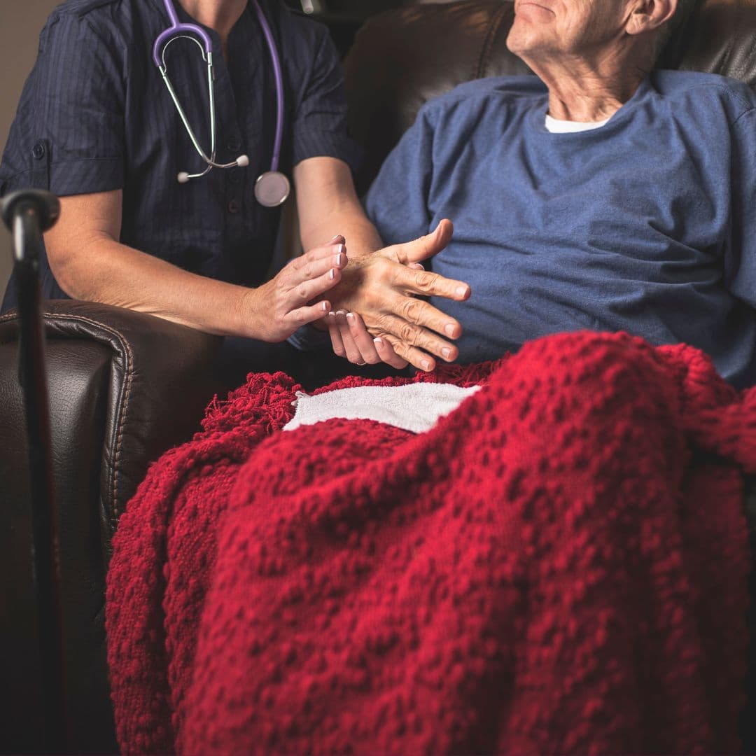 A caregiver comforts an elderly man in a chair covered with a red blanket, holding his hand and wearing a stethoscope. - Home Instead