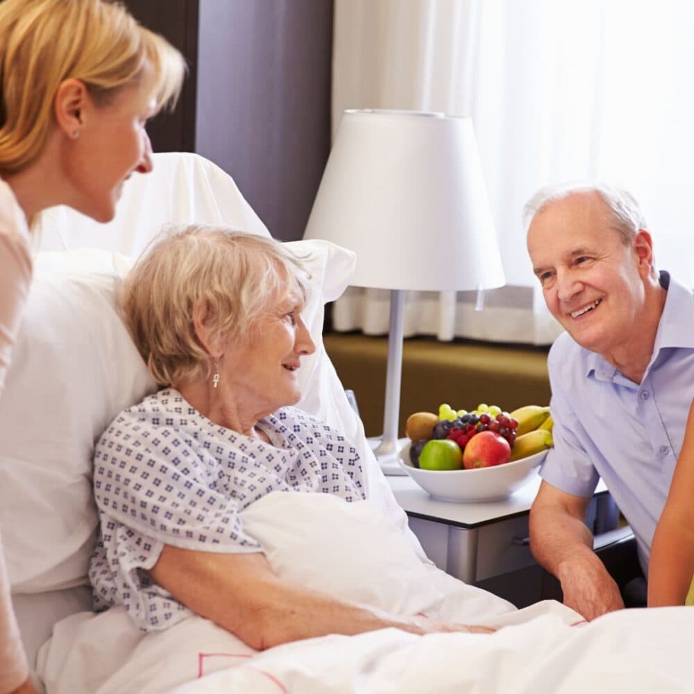 Two people visiting an elderly woman in a hospital bed, with a bedside table holding a bowl of fresh fruit. - Home Instead