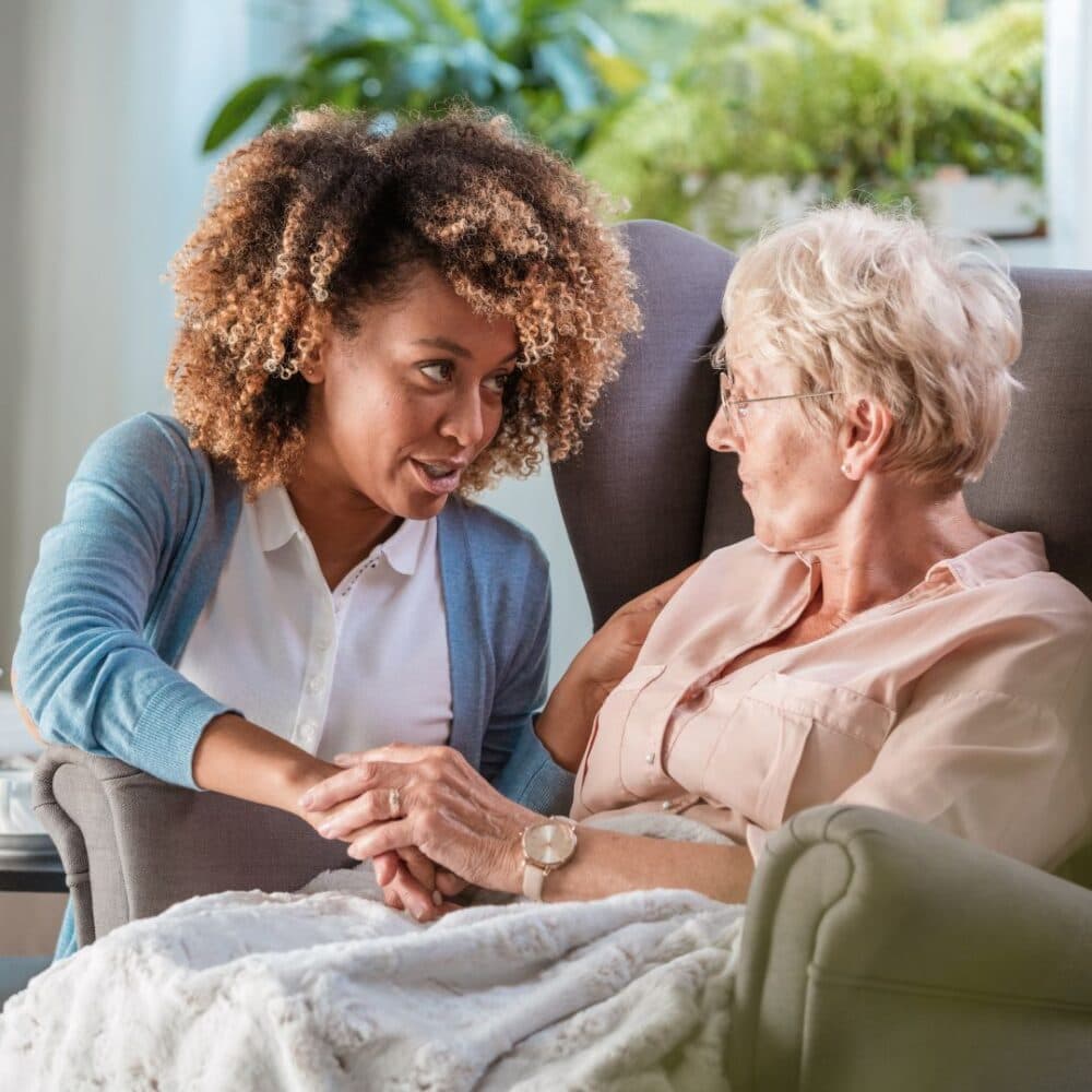 A younger woman with curly hair holds the hands of an older woman sitting in a chair, engaging in a caring conversation. - Home Instead