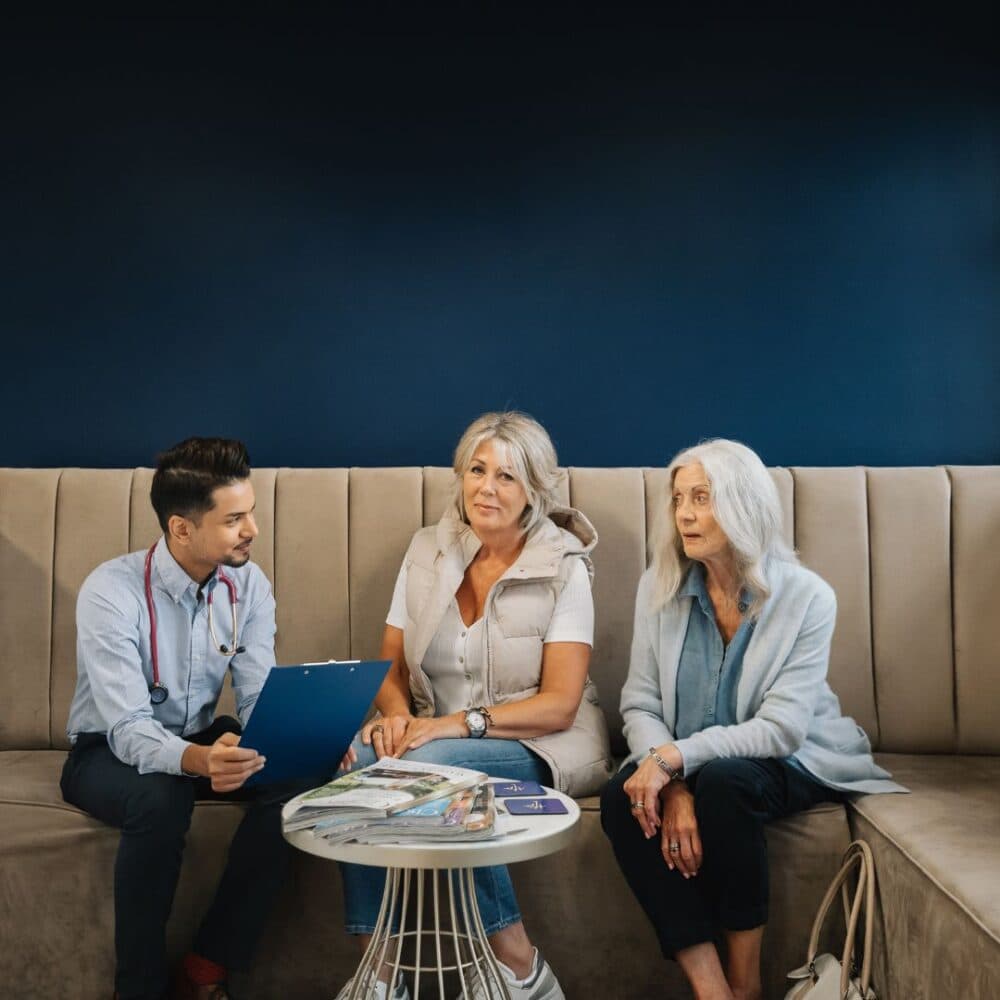 Doctor discussing with two women on a couch in a modern medical office, holding a clipboard with documents. - Home Instead
