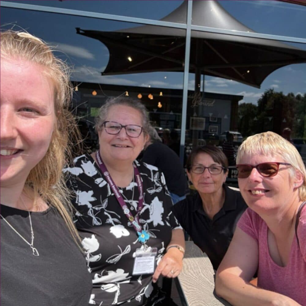 Four smiling people posing for a selfie outside a café with umbrellas and lights in the background. - Home Instead