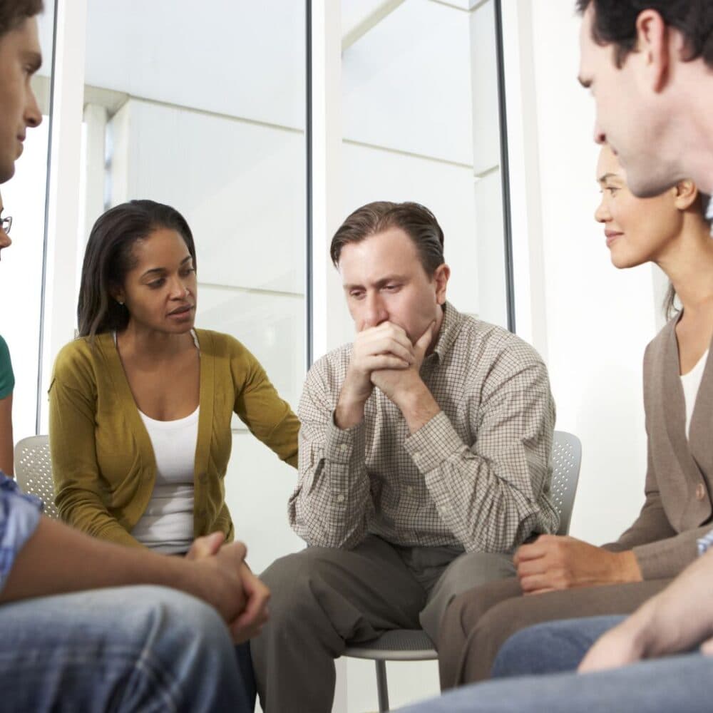 A group of people sitting in a circle, providing support to a man who looks distressed and is sitting with his hands clasped. - Home Instead