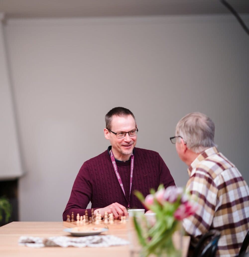 Two men are sitting at a table talking, with a chessboard and flowers in the foreground. - Home Instead
